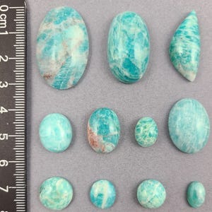May include: A collection of 11 smooth, polished, green and blue amazonite cabochons of varying sizes and shapes. The cabochons are arranged on a gray surface with a ruler for scale.
