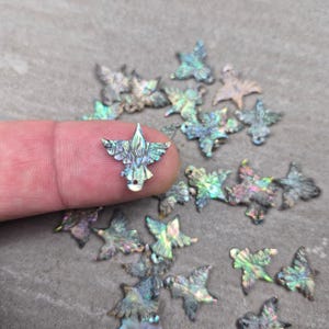 May include: Iridescent abalone shell charms in a leaf shape. The charms have a pearlescent sheen with colors of blue, green, and purple. A finger holds one charm, with several more scattered around.