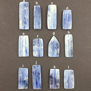 May include: Twelve blue kyanite stones, each with a unique shape and size, arranged in rows on a gray surface. The stones are translucent and have a striated pattern.