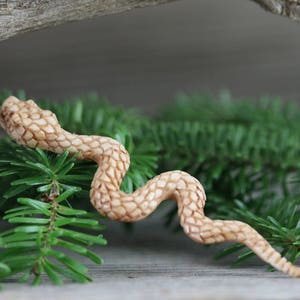 Puede incluir: Una figura de serpiente de madera tallada en color marr&oacute;n claro, posada sobre agujas de pino verdes. La serpiente tiene un patr&oacute;n texturizado y detallado y est&aacute; colocada en una pose curvada. El fondo es una superficie de madera borrosa.