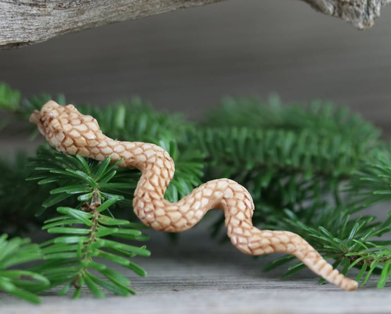 Puede incluir: Una figura de serpiente de madera tallada en color marr&oacute;n claro, posada sobre agujas de pino verdes. La serpiente tiene un patr&oacute;n texturizado y detallado y est&aacute; colocada en una pose curvada. El fondo es una superficie de madera borrosa.