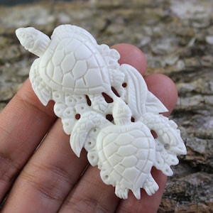 May include: Ivory-colored carved turtle figurine. The figurine features three turtles with detailed shell patterns and decorative elements. The turtles are arranged in a cluster, with intricate carvings around them. The overall design is detailed and artistic.
