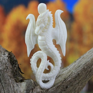 White hand-carved bone dragon pendant with wings and curled tail, side-drilled for hanging, shown on rustic wood.