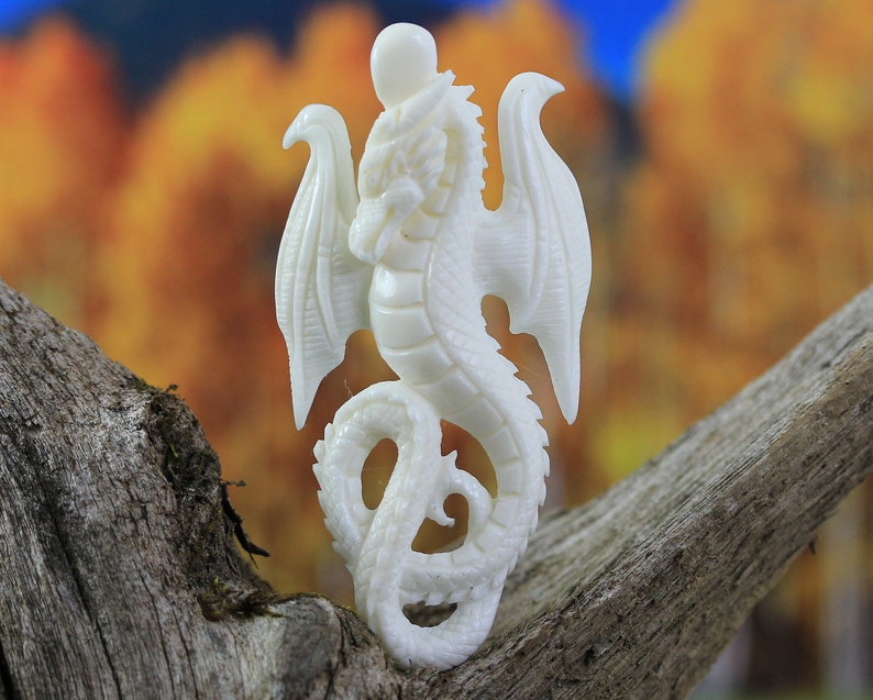 White hand-carved bone dragon pendant with wings and curled tail, side-drilled for hanging, shown on rustic wood.