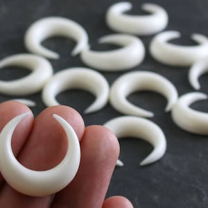 Ten small, white, crescent moon-shaped pieces, carved from buffalo bone, are scattered across a dark grey, textured background. One of the crescent moons is held between a thumb and forefinger, showing its size and detail against the skin.  Each piece has a smooth, matte finish and a subtle curve.
