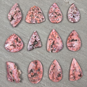 Natural ocean jasper cabochons with pink and coral orbicular patterns and natural druzy pockets arranged on a neutral stone background, each piece uniquely shaped.