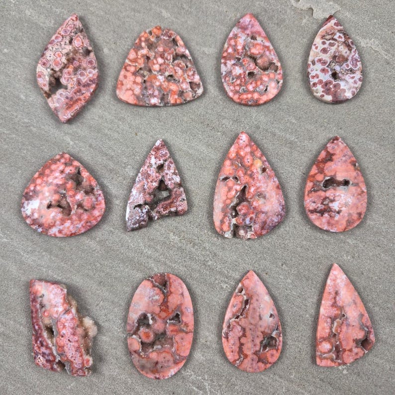 Natural ocean jasper cabochons with pink and coral orbicular patterns and natural druzy pockets arranged on a neutral stone background, each piece uniquely shaped.