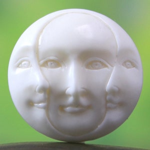 May include: A round, off-white button featuring three sculpted faces. The faces are arranged in a circular pattern, with each face partially overlapping the others. The button has a smooth, matte finish and is set against a blurred green background.