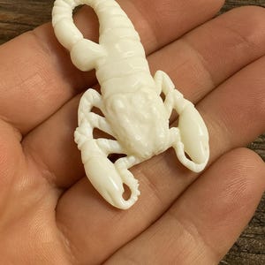 May include: A detailed, off-white scorpion figurine. The scorpion is sculpted with visible segments, claws, and a curved tail. The figurine is held in a hand, showcasing its size and intricate design. The overall theme is a decorative item or a piece of jewelry.