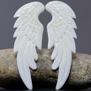 May include: Two ivory-colored, carved wing pendants. Each wing features detailed feather patterns and a curved shape. The wings are displayed against a neutral background, highlighting their intricate design and craftsmanship.