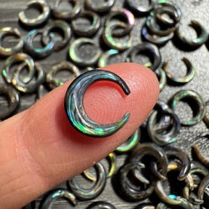 May include: Abalone shell crescent-shaped jewelry. The iridescent, curved piece is held by a finger, with many similar pieces in the background. The colors include blues, greens, and blacks, with a glossy finish. The jewelry is likely for ear piercings.