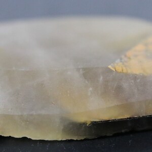 May include: A translucent, light gray, irregularly shaped stone with a thin, light brown layer on one side. The stone has a smooth, polished surface.