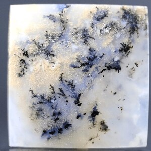 May include: Square-shaped gemstone with a dendritic pattern. The stone features a white base with blue and tan hues, and intricate black, tree-like inclusions. The polished surface reflects light, highlighting the natural patterns.