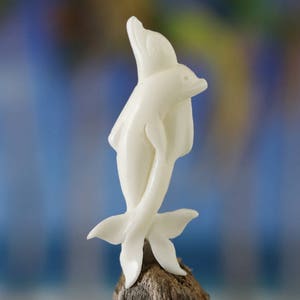 Könnte beinhalten: Eine weiße Skulptur von zwei Delfinen, die springen und ineinander verschlungen sind. Die Delfine sind aus einem hellfarbigen Material geschnitzt und auf einem Stück Treibholz montiert. Der Hintergrund ist ein verschwommenes Blau und Grün.