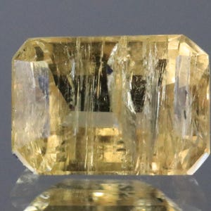 May include: A rectangular, faceted gemstone with a golden-yellow hue. The gem's interior displays visible inclusions and a reflective surface, suggesting a high degree of clarity. The cut of the stone is an emerald cut, with a polished appearance. The gem is likely a precious stone.
