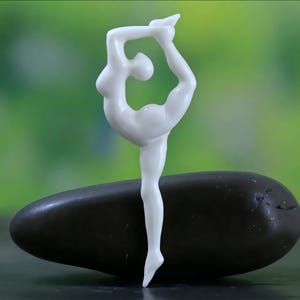Peut inclure: Une figurine en céramique blanche représentant une personne en posture de yoga, en équilibre sur une jambe et arquant le dos. La figurine est posée sur une pierre ovale gris foncé, sur fond vert flou.
