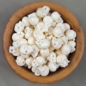 May include: A wooden bowl filled with small, white skull-shaped beads. The beads are clustered together, with visible eye sockets and other facial details. The bowl is made of light brown wood, and the background is a solid gray.