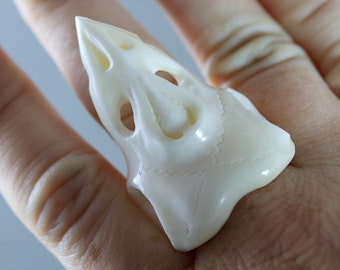 May include: A cream-colored, sculpted ring in the shape of a stylized skull. The ring features open eye sockets and other carved details, with a smooth, glossy finish. The ring is worn on a finger.