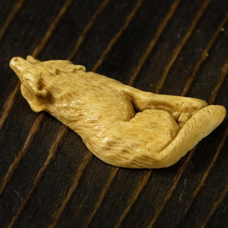 May include: A carved figurine of a wolf in a reclining pose. The wolf is a light tan color, with detailed fur texture. The figurine is lying on a dark brown, wood-grain surface.