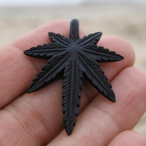 May include: A black, carved pendant in the shape of a cannabis leaf. The pendant has detailed veining and a loop at the top for a chain. The pendant is held in a person's hand.