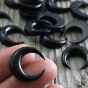 A close-up shot of several small, black, crescent moon-shaped pieces carved from bull horn, scattered across a light grey, wood-textured surface. One of the crescents is held between a thumb and forefinger, showing its size and detail against the wood grain. The crescents have a smooth, polished finish.