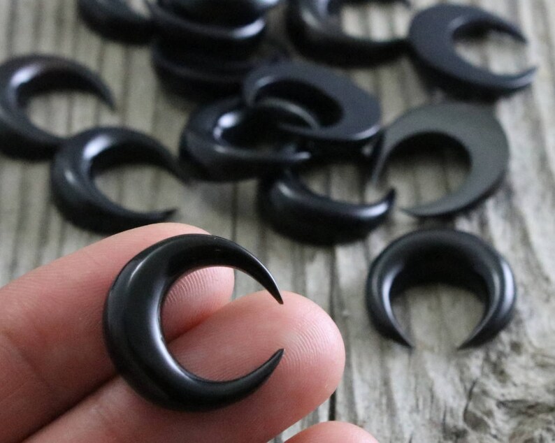 A close-up shot of several small, black, crescent moon-shaped pieces carved from bull horn, scattered across a light grey, wood-textured surface. One of the crescents is held between a thumb and forefinger, showing its size and detail against the wood grain. The crescents have a smooth, polished finish.