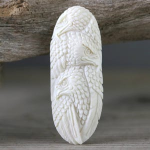 May include: An oval-shaped off-white pendant featuring a detailed relief carving of three eagle heads stacked vertically. The surface displays intricate feather textures and sharp, defined beaks, creating a layered, sculptural appearance.