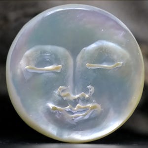 May include: A white, round, iridescent moon pendant with a carved face. The face has closed eyes and a slightly smiling mouth.