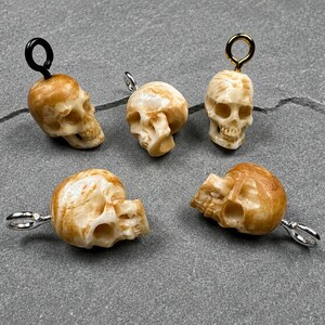 May include: Five skull-shaped pendants with a beige and tan marbled finish. Each pendant features a metal jump ring at the top, including one black, one gold-toned, and three silver-toned rings, arranged in a group on a gray textured surface.