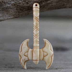 May include: A carved, off-white axe pendant with a detailed handle and double-bladed head. The handle features a crisscross pattern, and the axe head has a symmetrical design. The pendant is likely made of bone or a similar material.