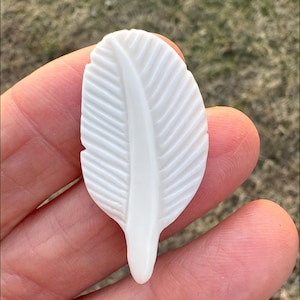 Puede incluir: Un colgante de pluma tallada, de color blanco roto. La pluma tiene vetas detalladas y un tallo central liso. El colgante se sostiene en una mano, mostrando su delicado diseño y textura. La pluma mide aproximadamente 7.6 cm de largo.