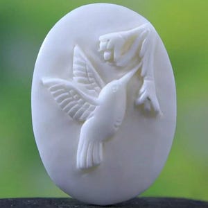 Op de afbeelding: Ovale, crèmekleurige gravure met een kolibrie in vlucht, die naar een bloem reikt. De vleugels van de vogel zijn uitgespreid en zijn snavel is uitgestrekt naar de bloem. De achtergrond is een zacht, wazig groen.