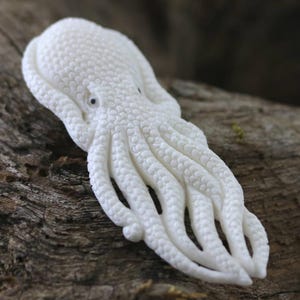 May include: A detailed, white octopus sculpture with textured skin and eight tentacles. The sculpture is placed on a piece of weathered wood, showcasing the intricate details of the sea creature.