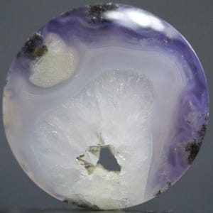 Può includere: Una gemma rotonda e lucida con un motivo sorprendente. La pietra presenta bande concentriche bianche e lavanda, con una formazione cristallina centrale e una piccola cavità. Macchie scure sono sparse sul bordo.