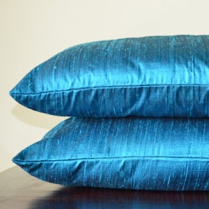 May include: Two teal-colored throw pillows stacked on top of each other. The pillows have a rectangular shape and are made of a shiny, textured fabric. The pillows are a vibrant shade of teal, adding a pop of color to any room.