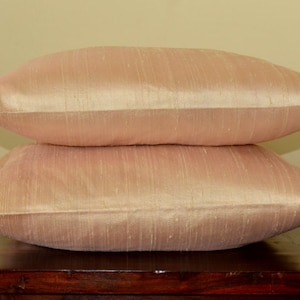 May include: Two peach-colored pillow covers made of silk. The pillow covers are stacked on top of each other on a dark brown wooden surface.