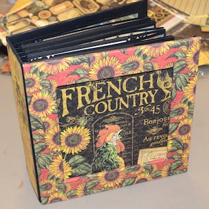 Puede incluir: Un álbum negro con un diseño de girasol y gallo. La portada dice "French Country No. 45" con el texto "Bonjour" y "Au revoir goodbye" en una fuente más pequeña.