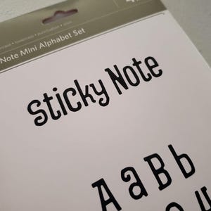Può includere: Un set di mini note adesive con alfabeto in una confezione con sfondo bianco e testo nero. L'intestazione recita Sticky Note Mini Alphabet Set, e il sottotesto indica che include maiuscole, minuscole, punteggiatura e simboli.