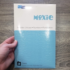 Puede incluir: Un paquete para un juego de alfabeto Unicase Moxie clásico. El paquete es blanco y azul claro con la palabra "Moxie" en letras grandes de color azul claro. Incluye Unicase, números y puntuación.
