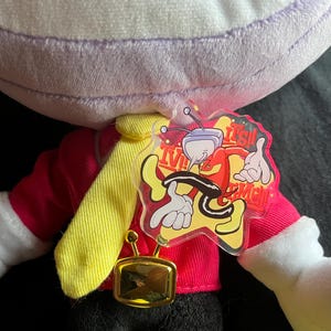 May include: A plush doll with a purple head, red jacket, yellow tie, and black pants. The doll has white gloves and a gold-colored rectangular pendant. A clear tag with a cartoon character and the text "It's Me!" is attached.