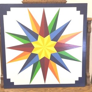 May include: A square, colorful starburst design on a white background, framed by a dark blue border. The star features vibrant hues of yellow, red, orange, green, and blue, creating a striking visual effect. The artwork is likely a decorative piece.