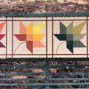 May include: A rectangular wooden sign featuring four square panels, each displaying a stylized leaf design. The leaves are rendered in shades of red, orange, yellow, green, and blue. The sign is painted with a neutral background and brown borders.