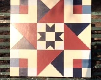 May include: Square wooden quilt block painted with a geometric design. The quilt block features a central star pattern surrounded by triangles and squares in red, navy, cream, and gray. The block is displayed outdoors on a green bench.