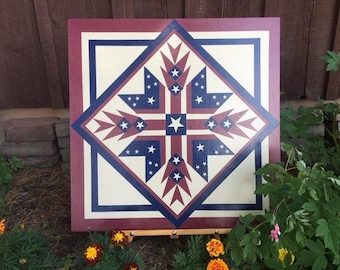 May include: A red, white, and blue painted wooden barn quilt with a star pattern. The quilt is on an easel and is in front of a brick wall and green plants.