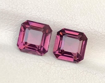 May include: Two square-cut, deep pink gemstones with a lighter pink center. The gems are set against a white background, likely in a jewelry setting. The gems have a faceted cut, reflecting light.