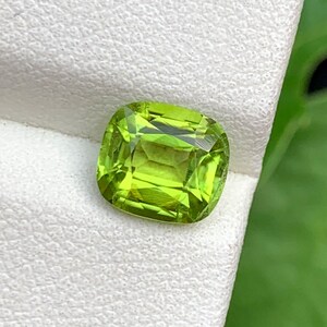 Op de afbeelding: Een levendige, kussenvormig geslepen peridot edelsteen. De edelsteen heeft een heldere, limoengroene tint met facetten die het licht vangen. De steen is geplaatst tegen een witte achtergrond, met een vage groene achtergrond.