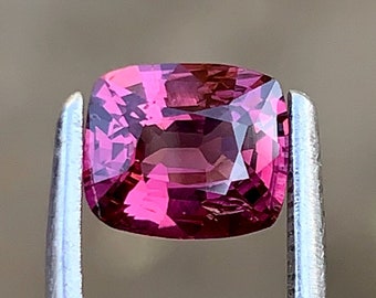 Op de afbeelding: Een kussenvormige edelsteen, vastgehouden door een pincet. De edelsteen is diep roze van kleur met facetten die het licht vangen. De edelsteen heeft een vierkante vorm met afgeronde hoeken.