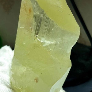 May include: A translucent, pale yellow crystal with a rectangular shape and a rough, uneven edge. The crystal has visible internal striations and is held by a white glove. The overall appearance is of a natural mineral specimen.