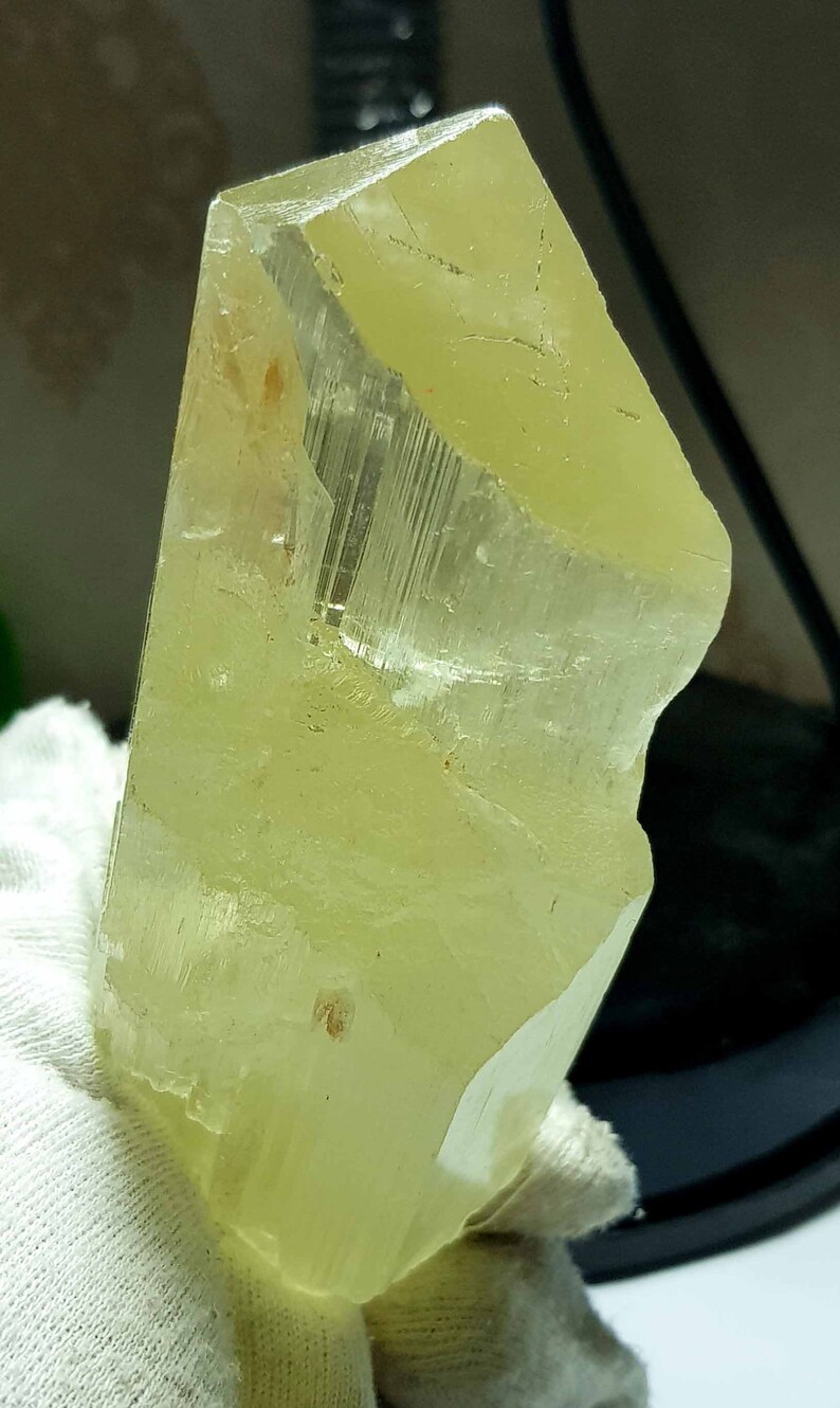 May include: A translucent, pale yellow crystal with a rectangular shape and a rough, uneven edge. The crystal has visible internal striations and is held by a white glove. The overall appearance is of a natural mineral specimen.