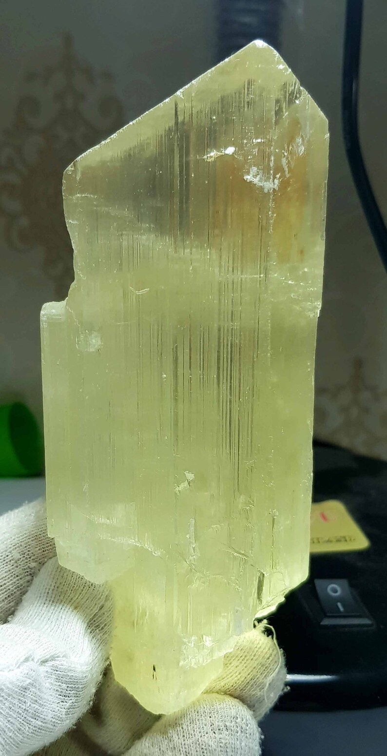 May include: A translucent, pale yellow crystal formation. The crystal has a rectangular shape with a rough, textured surface and visible internal striations. The overall color is a soft, light yellow.